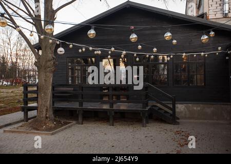 Retro Girlande Lampen draußen leuchten am Abend an den Wänden des schwarzen Hauses. Hinterhof Beleuchtung, Licht im Abendgarten, elektrische Laterne Stockfoto