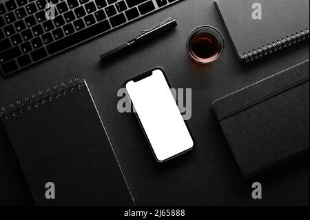 Overhead Shot, moderne schwarze Arbeitsplatzkompositionen mit Tastatur, Notebook, Stift, Kaffee und Smartphone-Modell mit weißem Bildschirm auf schwarzem Hintergrund. Stockfoto