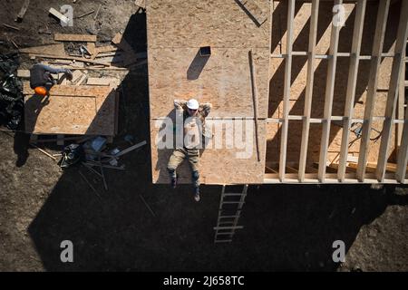 Porträt eines männlichen Arbeiters, der ein Holzhaus baute. Mann, der auf dem Dach des Gebäudes auf der Baustelle in weißem Helm lag. Drohnenansicht Stockfoto