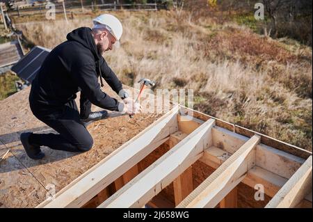 Zimmermann Hämmerschläge Nagel in die OSB-Platte auf dem Dach der zukünftigen Hütte. Mann Arbeiter Gebäude Holzrahmen Haus. Zimmerei- und Konstruktionskonzept. Stockfoto