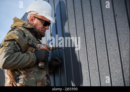 Männlicher Baumeister, der schwarze Wellblech installiert, das als Fassade des zukünftigen Häuschens verwendet wird. Mann Arbeiter Gebäude Holzrahmen Haus. Zimmerei- und Konstruktionskonzept. Stockfoto