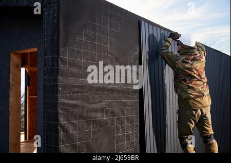 Männlicher Baumeister, der schwarze Wellblech installiert, das als Fassade des zukünftigen Häuschens verwendet wird. Mann Arbeiter Gebäude Holzrahmen Haus. Zimmerei- und Konstruktionskonzept. Stockfoto