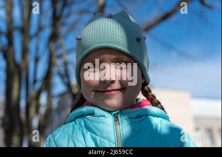 Ein Porträt eines glücklichen ukrainischen Mädchens gegen den blauen Himmel und die Bäume. Ein fünfjähriges Kind lächelt ein schlaues Lächeln, während sie an der Kamera vorbeischaut. Spring tim Stockfoto
