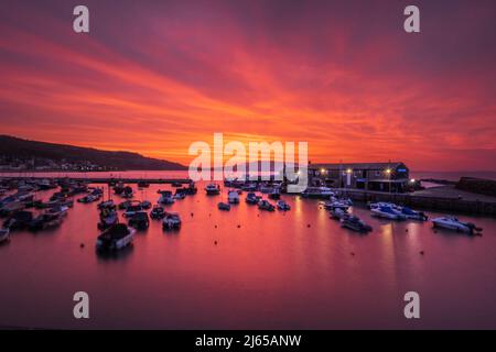 Lyme Regis, Großbritannien. 28. April 2022. Der Morgenhimmel über dem Cobb Habour bei Lyme Regis schillert bei Sonnenaufgang mit leuchtendem Rot und Orange. Kredit: Celia McMahon/Alamy Live Nachrichten Stockfoto