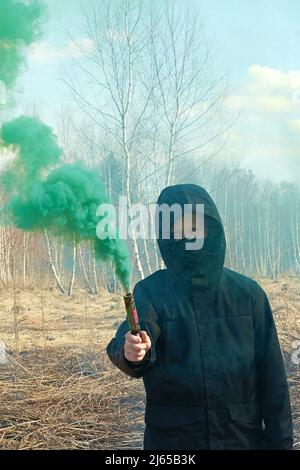 Ein Teenager in einer schwarzen Jacke und einer Maske hält eine grüne Rauchbombe Stockfoto