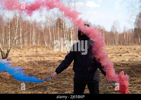 Ein Teenager in einer schwarzen Jacke und einer Maske hält zwei farbige Rauchbomben Stockfoto