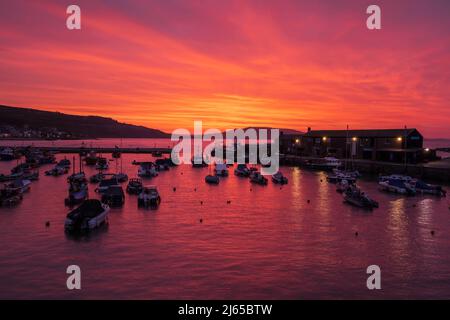 Lyme Regis, Großbritannien. 28. April 2022. Der Morgenhimmel über dem Cobb Habour bei Lyme Regis schillert bei Sonnenaufgang mit leuchtendem Rot und Orange. Kredit: Celia McMahon/Alamy Live Nachrichten Stockfoto