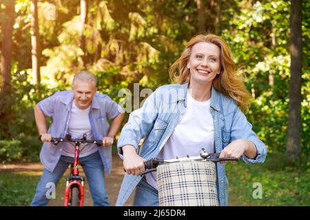Fröhliches aktives Seniorenpaar mit Fahrrad, das gemeinsam durch den Park läuft. Perfekte Aktivitäten für Senioren im Ruhestand Lebensstil tragen Casual tragen tragen lächelnd Stockfoto