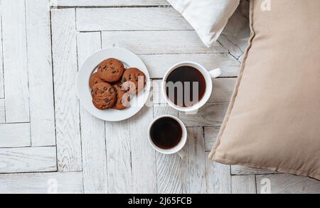 Frühstück am Morgen. Zwei weiße Tassen Kaffee und Schokoladenkekse stehen auf einem hellen Holzparkett und Kissen liegen in der Nähe. Gemütlichkeit, Tag Stockfoto