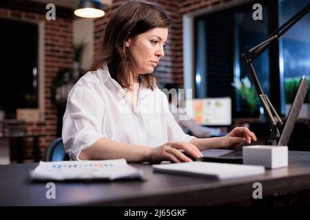 Geschäftsfrau des Marketingunternehmens, die im Büroarbeitsplatz sitzt, während sie die Buchhaltungsunterlagen durchgeht. Starten Sie den Projektmanager mithilfe eines Laptops, um Finanzdiagramme zu analysieren und das Kampagnenbudget zu fördern. Stockfoto