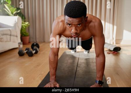 Afroamerikanischer junger Mann mit hoher Planke in seinem warm beleuchteten Wohnzimmer, der zu Hause arbeitet und dabei ist, einen Push-up zu machen Stockfoto