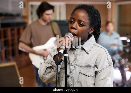 Waist-up-Porträt einer jungen schwarzen Frau, die zum Mikrofon singt, während sie im professionellen Studio Musik mit Band aufnimmt Stockfoto