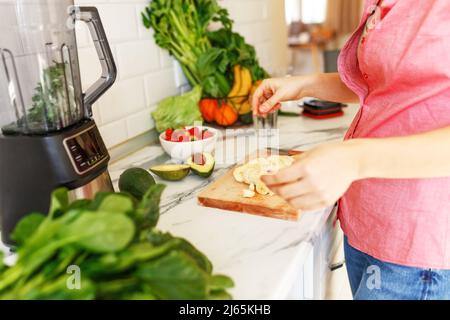 Eine Frau, die mit dem Mixer Obst-Smoothies macht. Gesunde Ernährung, Ernährung Lifestyle-Konzept Porträt einer jungen Frau, die zu Hause in der Küche ein Getränk mit Bananen, Avocado und Spinat zubereitet. Stockfoto