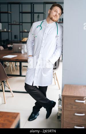 Vertikales Porträt eines träumenden männlichen Arztes in weißer medizinischer Uniform, stehend mit Stethoskop in der Nähe des Fensters im Krankenhausbüro und schauend nach oben. Stockfoto