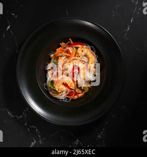 Funchoza mit Garnelen und Gemüse auf schwarzem Hintergrund, Kopierplatz. Glasnudeln, traditionelle asiatische Speisen, Braten Sie Nudeln oder Wok-Nudeln in einer bla Stockfoto