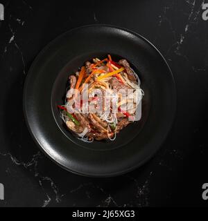 Funchoza mit Rindfleisch und Gemüse auf schwarzem Hintergrund, Platz zum Kopieren. Glasnudeln, traditionelle asiatische Speisen, Braten Sie Nudeln oder Wok-Nudeln in einem schwarzen Stockfoto