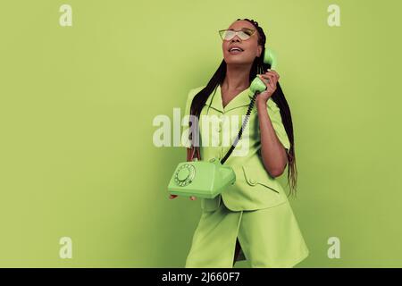 Monochromes Porträt eines lächelnden afrikanischen Mädchens trägt Retro-Stil Outfit halten vintage Telefon isoliert auf grünem Hintergrund. Konzept der Emotionen, Schönheit Stockfoto