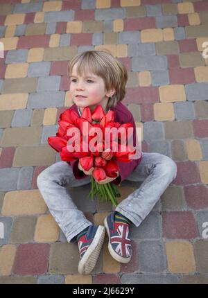 Netter Vorschuljunge sitzt auf einer Pflasterplatte mit einem großen Bouquet aus roten Tulpen. Ein Geschenk mit Liebe zu Mama, Großmutter. Muttertag. Dankbarkeit. Frühlingsmeere Stockfoto