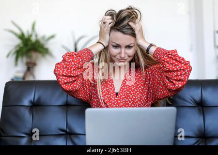Die junge blonde Frau, die zu Hause einen Laptop benutzt, sitzt auf dem Sofa und ist mit den Händen am Kopf verängstigt und erstaunt Stockfoto