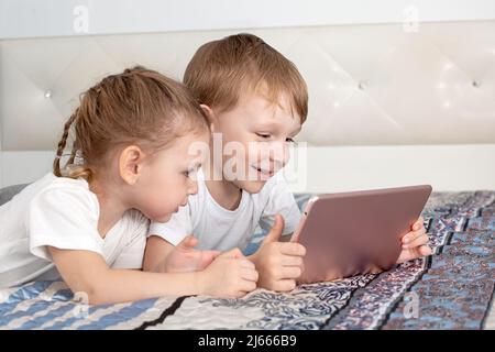Kinder und Gadgets. Ein Junge und ein Mädchen liegen auf dem Bett, schauen auf die Tafel und lächeln Stockfoto