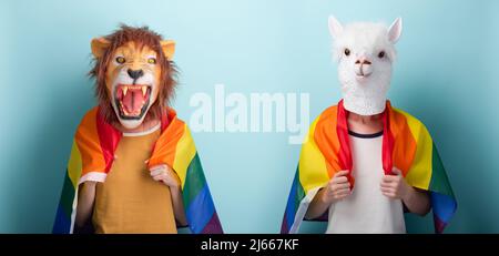 Junge Frau in Löwen- und Alpakamaske hält eine lgbt-Stolzflagge in Regenbogenfarben, die auf blauem Hintergrund isoliert um die Schultern gewickelt ist Stockfoto