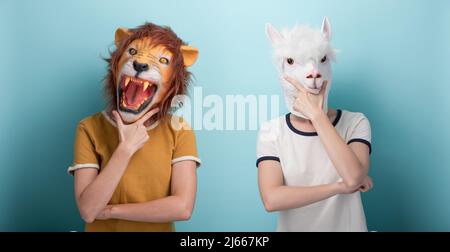 Junge Frau in Löwenmaske und Alpaka-Maske mit Hand am Kinn, denken und Entscheidungen treffen, nachdenklicher Ausdruck, isoliert auf blauem Hintergrund Stockfoto