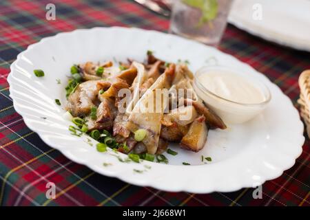 Köstliche geräucherte Schweinöhren, serviert mit saurer Rahm und Kräutern auf einem weißen Teller in einem Restaurant Stockfoto