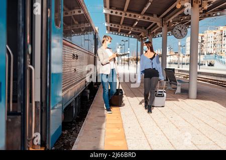 Zwei glückliche Freundinnen in Schutzmasken gehen und plaudern auf dem Bahnsteig am Bahnhof Stockfoto