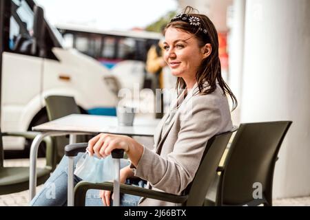 Eine junge Reisende mit dem Gepäck und der Schutzmaske sitzt im Café und wartet auf ihren Bus zu kommen Stockfoto