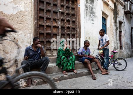 Menschen sitzen vor alter Tuere in Gasse, Stone Town, UNESCO Weltkulturerbe, Altstadt, Sansibar, Unguja, Tansania Stockfoto