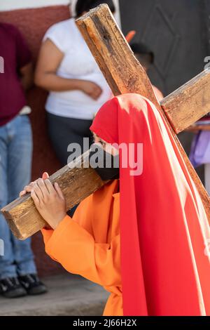Eine junge Mexikanerin trägt in Kostümen ein Holzkreuz durch die Straßen während einer Prozession zum Karsamstag von der St. Francisco Assisi Kirche am 16. April 2022 in Patzcuaro, Michoacan, Mexiko. Die kleine indigene Stadt bewahrt die Traditionen der spanischen Kolonialherrschaft, einschließlich der Bruderschaft der Büßer während der Karwoche. Stockfoto
