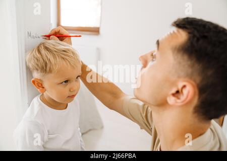Weißer Mann, der zu Hause die Körpergröße seines Sohnes misst und auf Türrahmen zeichnet Stockfoto