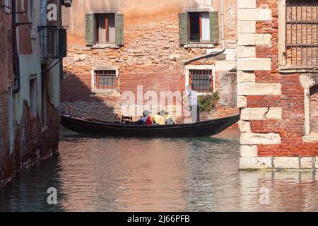 Ein venezianischer Gondolier in einer traditionellen schwarzen Gondel schwebt über einem sonnendurchfluteten Kanal. Stockfoto