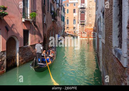 Ein venezianischer Gondolier in einer traditionellen schwarzen Gondel schwebt über einem sonnendurchfluteten Kanal. Stockfoto