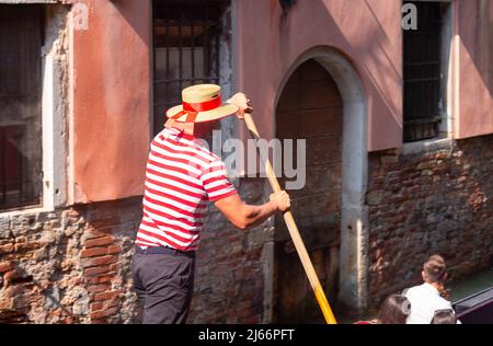 Ein venezianischer Gondolier in einer traditionellen schwarzen Gondel schwebt über einem sonnendurchfluteten Kanal. Stockfoto