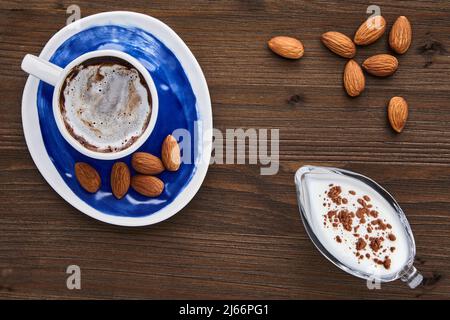 Tasse Cappuccino-Mandeln und Milch in einem Milchkännchen aus Glas auf einem dunklen Holztisch Stockfoto