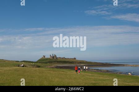 Landschaftsbild des fernen Dunstanburgh Castle auf der Whinsill vom Küstenpfad aus gesehen von Craster mit Menschen, die den Weg entlang gehen Stockfoto