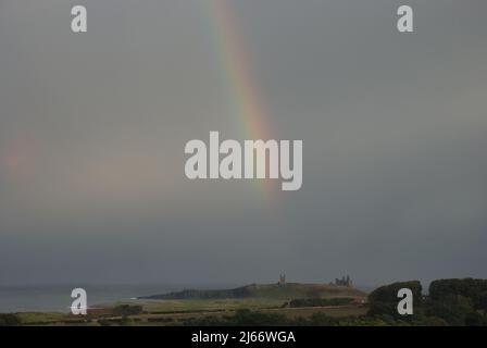 Landschaft an der Küste von Northumberland mit Dunstanburgh Castle in der Ferne und Regenbogen, der vor dunklem Himmel herabrast Stockfoto