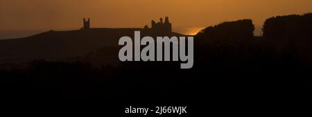 Blick auf das ferne Dunstanburgh Castle bei Sonnenuntergang mit einem Schloss in Silhouette vor einem orangenen Himmel und einem golden getippten Meer Stockfoto