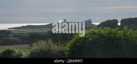 Eine ferne Burg von Dunstanburgh, die über Ackerland und gegen den Himmel gesehen wird, mit Sonnenlicht, das die Meeresoberfläche dahinter auffängt und beleuchtet Stockfoto