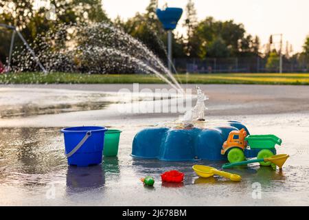 Spielplatz für Kinder im öffentlichen Park im Sommer ohne Menschen. Brunnen mit Spritzwasser und ließ Kinderspielzeug auf dem Boden auf einem sonnigen Stockfoto