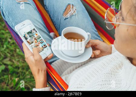 Pretty Content Creator in der Hängematte verwenden Smartphone Blick auf Bildschirm-Chat in sozialen Medien trinken Tee lesen Nachrichten online Browsing Bild im Urlaub, Rückansicht Stockfoto