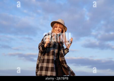 Junge kaukasische Mädchen hält Smartphone und Kreditkarte glücklich, verrückt oder erstaunt für den Erfolg. Aufgeregt Hipster Tourist Frau trägt Hemd und Hut macht schockiert Sieger Geste im Freien mit Himmel Hintergrund Stockfoto