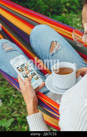 Bloggerin lesen Kommentar auf Smartphone-Gerät Tee trinken Entspannen Sie sich in der Hängematte im Herbst Sommer Frühling.Frau, die auf Handy-Bildschirm im Internet surfen Stockfoto