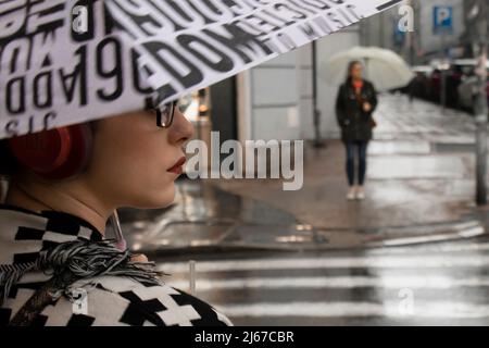 Belgrad, Serbien - 27. April 2022: Junge Frau im schwarz-weißen Muster-Outfit mit roten Kopfhörern, rotem Lippenstift und Brillen unter Regenschirm Stockfoto