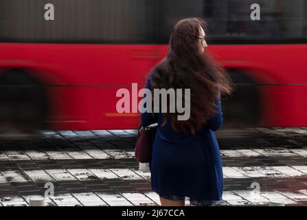 Belgrad, Serbien - 27. April 2022: Junge Frau mit langen, welligen Haaren wartet an einem regnerischen Tag auf einer Kreuzung mit verwacklungsunscharfem Verkehr, Rückansicht Stockfoto