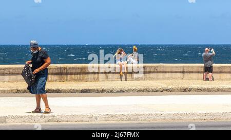 Menschen in der Ufermauer von El Malecon. Eine ältere kubanische Frau geht in den Avenue Divider. Zwei junge Frauen sitzen in der Wand, während ein Tourist Fotos macht Stockfoto