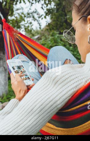 Bloggerin lesen Kommentar auf Smartphone-Gerät Tee trinken Entspannen Sie sich in der Hängematte im Herbst Sommer Frühling.Frau, die auf Handy-Bildschirm im Internet surfen Stockfoto