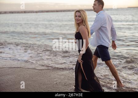 Attraktive Frau in schwarzem Kleid zu Fuß am Sandstrand mit Mann Stockfoto