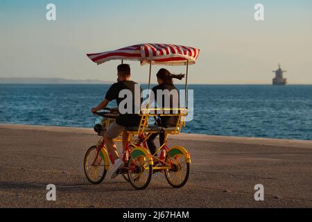 Junges Paar bei Sonnenuntergang auf einem vierrädrigen Fahrrad (surrey Bike) an der Promenade in der Nähe des Meeres. Stockfoto